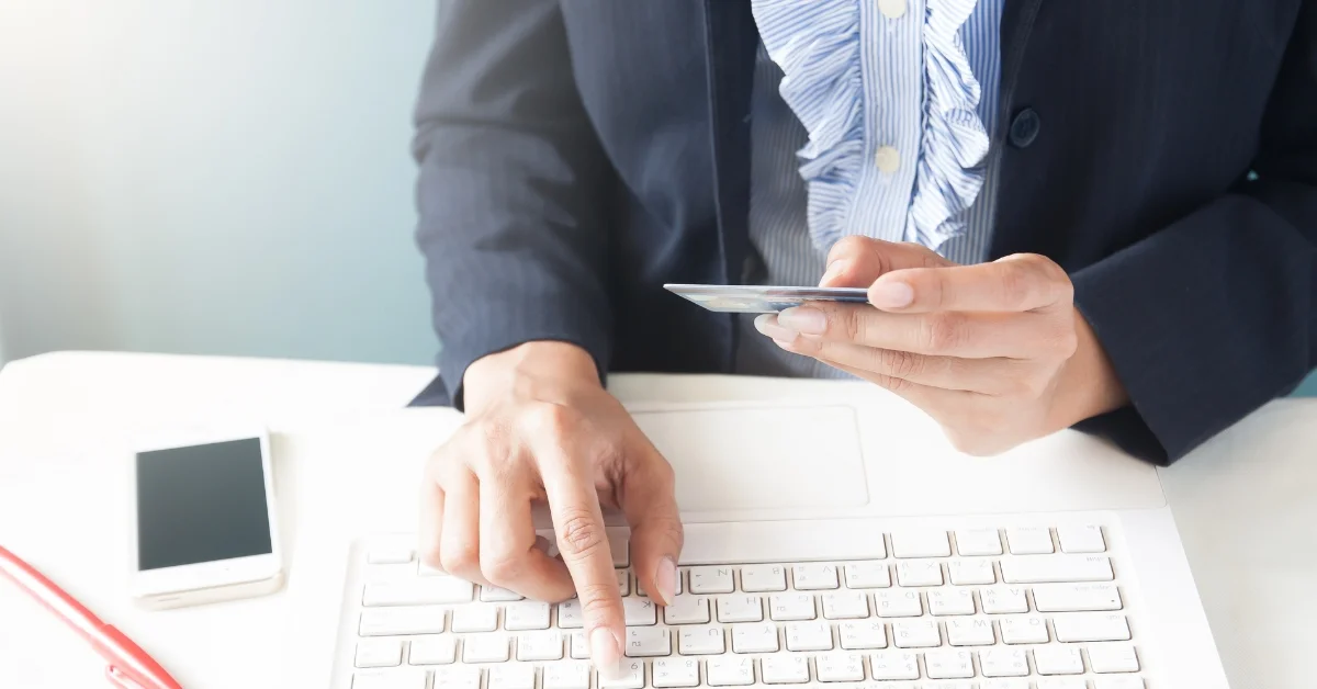 Image of A businesswoman in a dark blazer and ruffled blouse is sitting at a desk, typing on a white laptop with one hand while holding a bank card in the other. A smartphone is resting on the desk nearby.